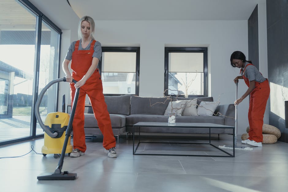 Two professional cleaners from Greenwich Cleaner stand back-to-back against a plain white wall, dressed in beige uniforms. The cleaner on the left, with dark hair pulled back, holds a vacuum cleaner w