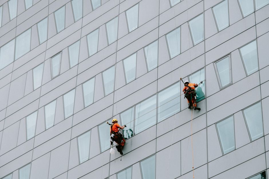 Two professional window cleaners wearing red safety helmets, harnesses, and gloves are suspended on ropes cleaning the exterior glass panels of a modern office building. One cleaner, on the left, is u