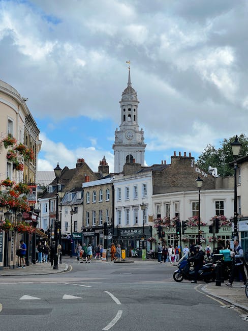 A commercial window cleaner wearing a black beanie, grey hoodie, and blue jeans is using a squeegee attached to an extension pole to clean large glass windows of a building on Greenwich High Road. The