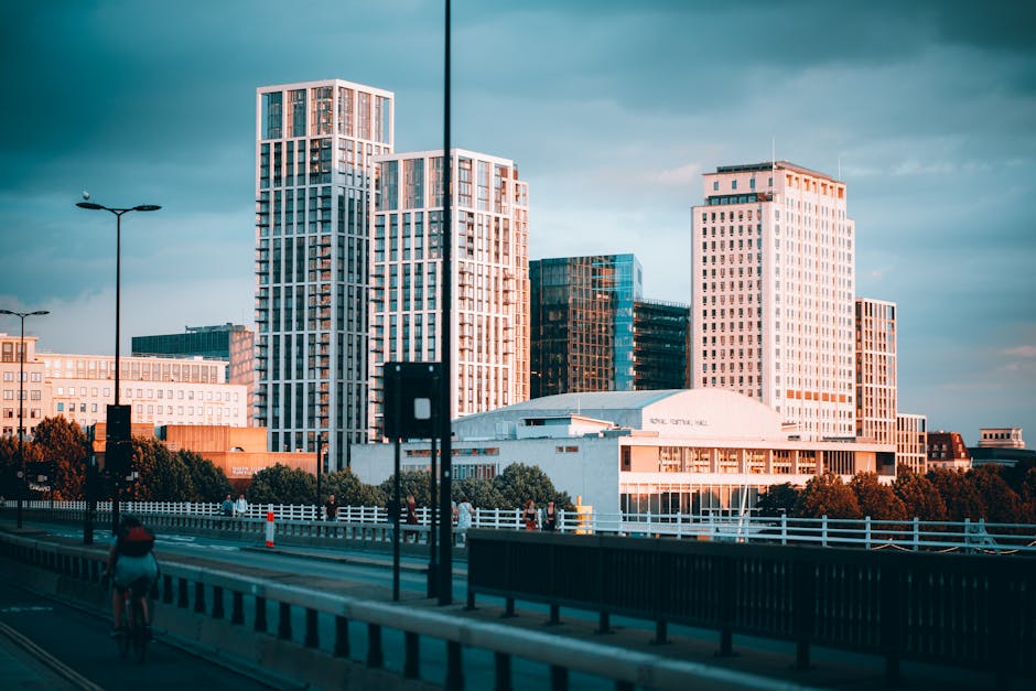 A daytime cityscape featuring the O2 Arena in Greenwich, with its distinctive air-supported roof structure and tall masts leaning at various angles, set against a cloudy sky. The image shows modern hi